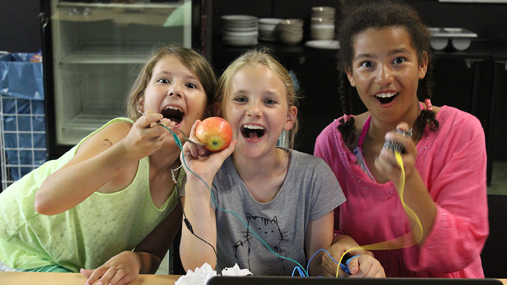 Drei Mädchen halten Apfel und Kabel in die Kamera und lachen