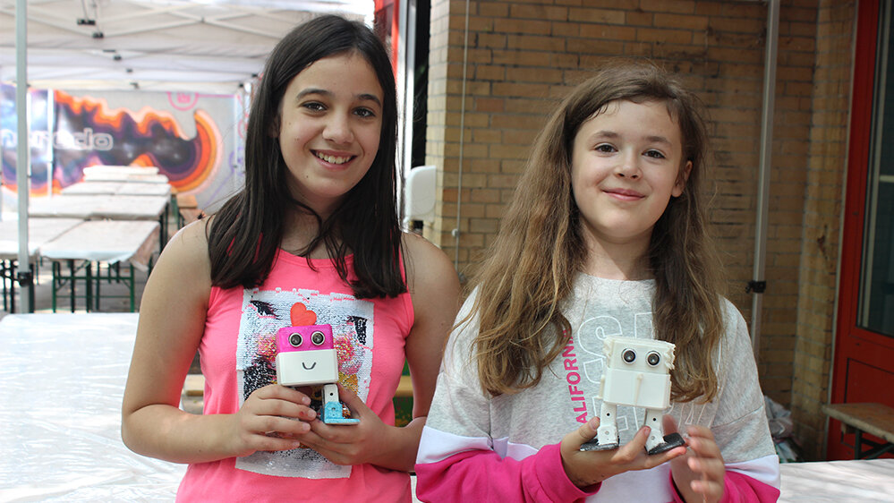 zwei Mädchen halten je einen selbstgebauten Roboter und lächeln