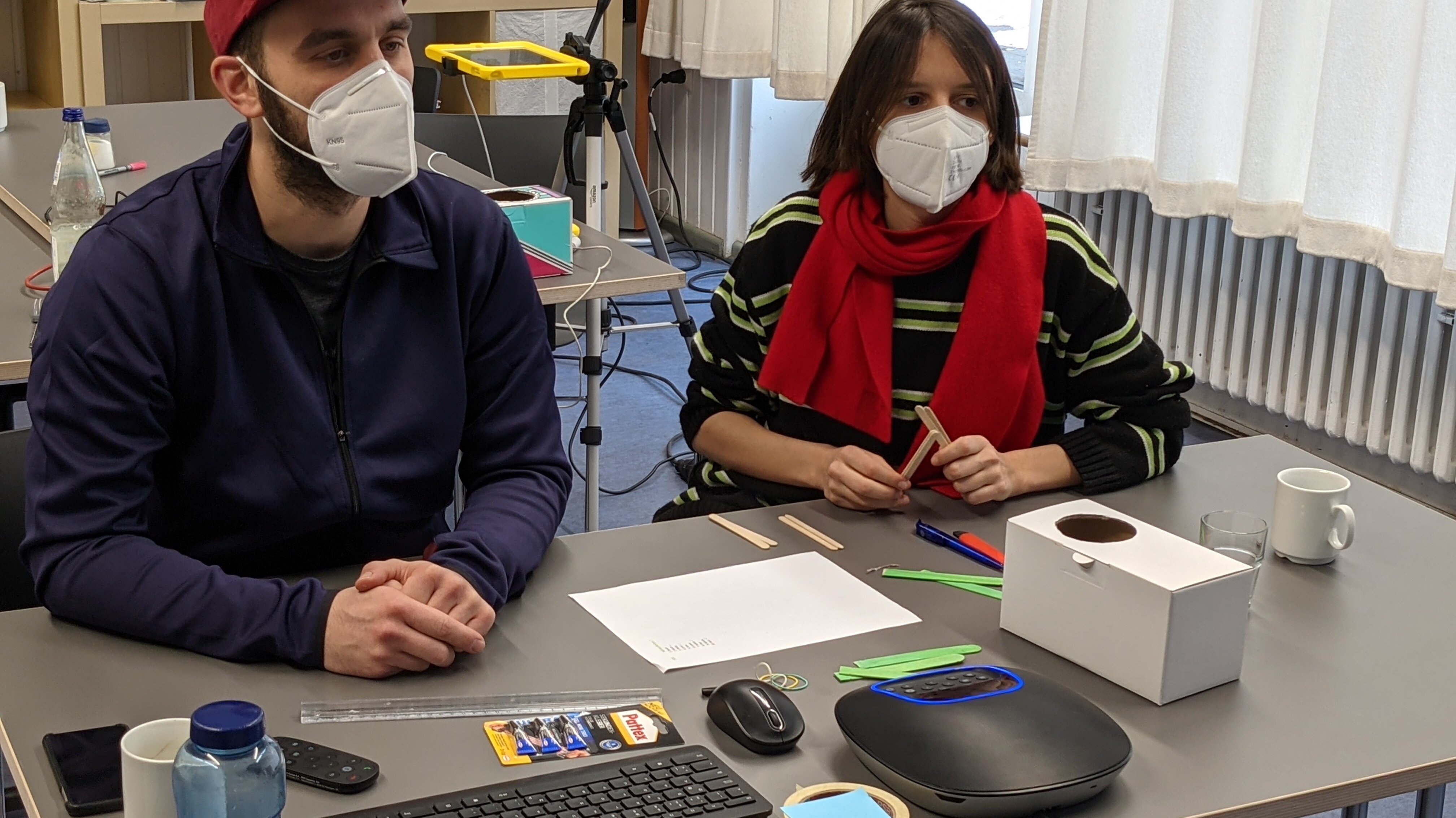 Zwei erwachsene Personen mit Maske sitzen an einem Tisch mit Technik