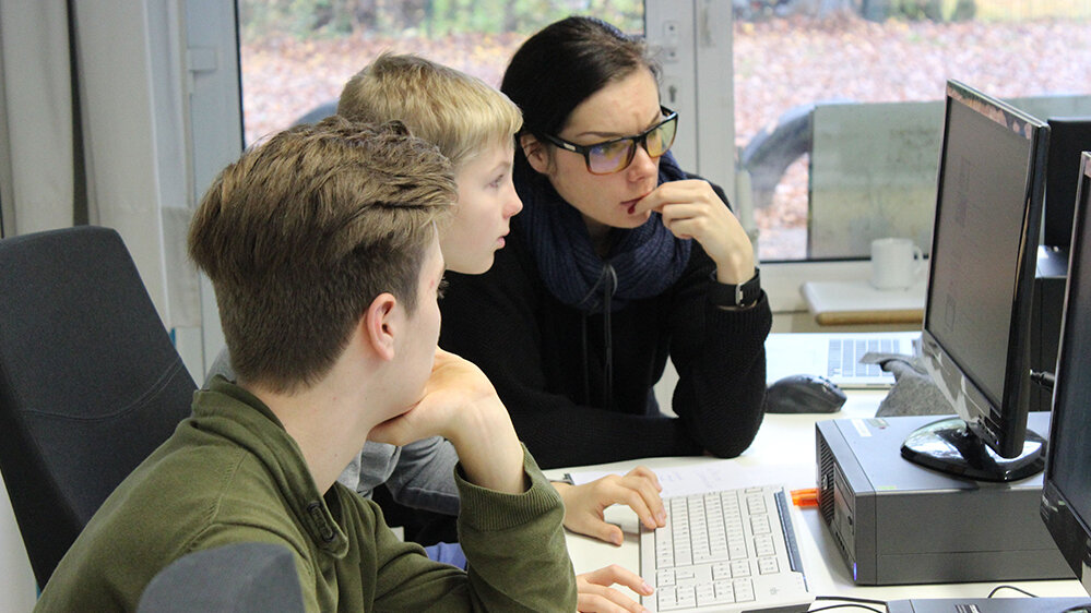 Zwei Jugendliche sitzen mit einer Dozentin am Tisch und schauen auf einen Computerbildschirm.