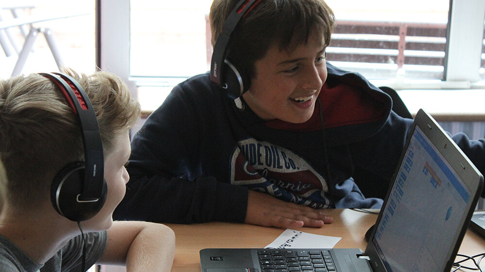 Zwei Jungen sitzen am Tisch mit einem Laptop und lachen.