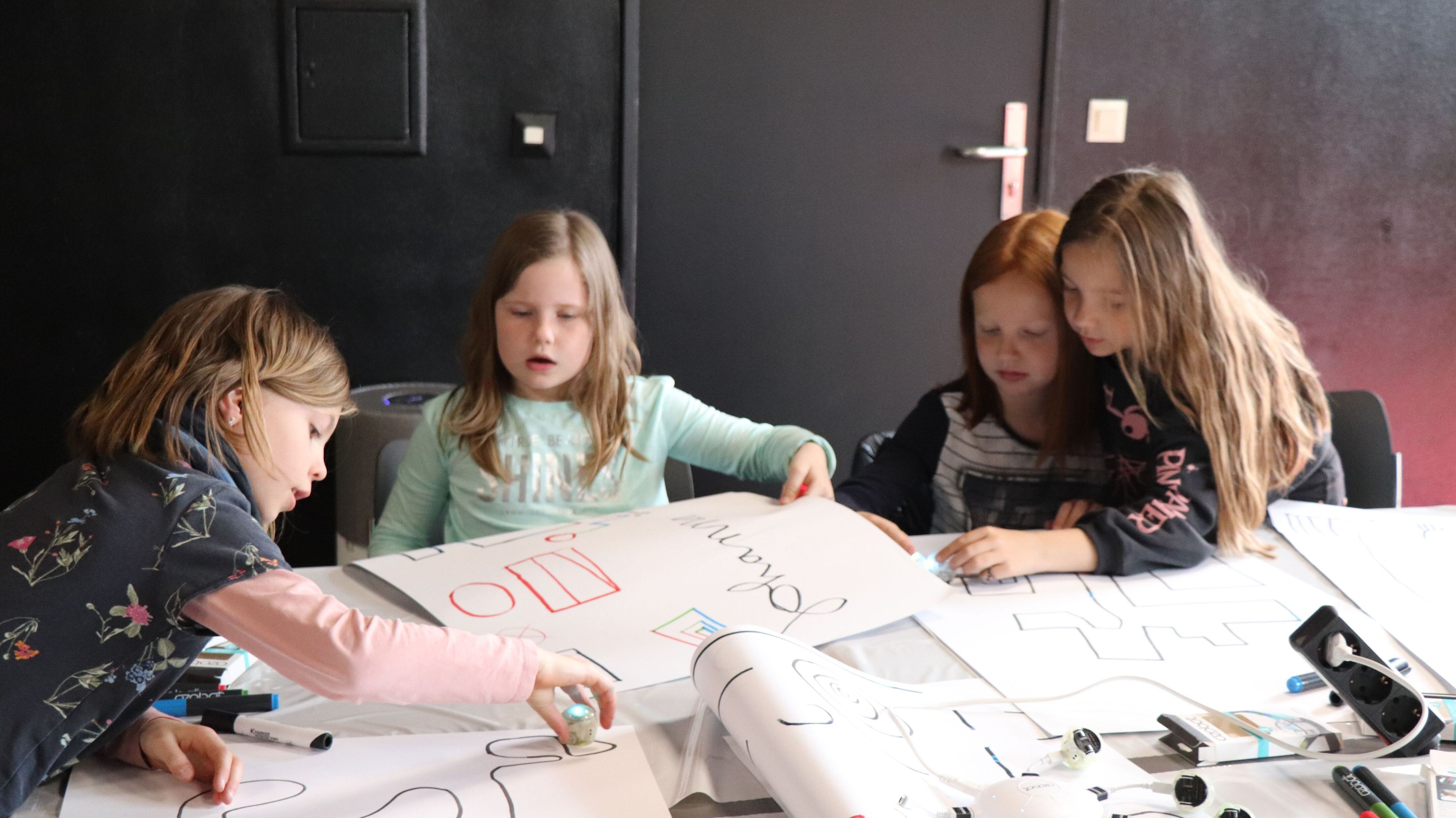 vier Mädchen sitzen an einem Tisch und programmieren Roboter mit Farbstiften