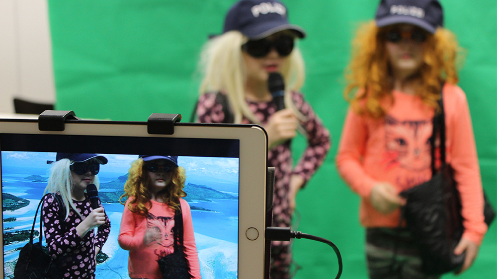Zwei Mädchen werden vor einem grünen Stoff von einem iPad gefilmt