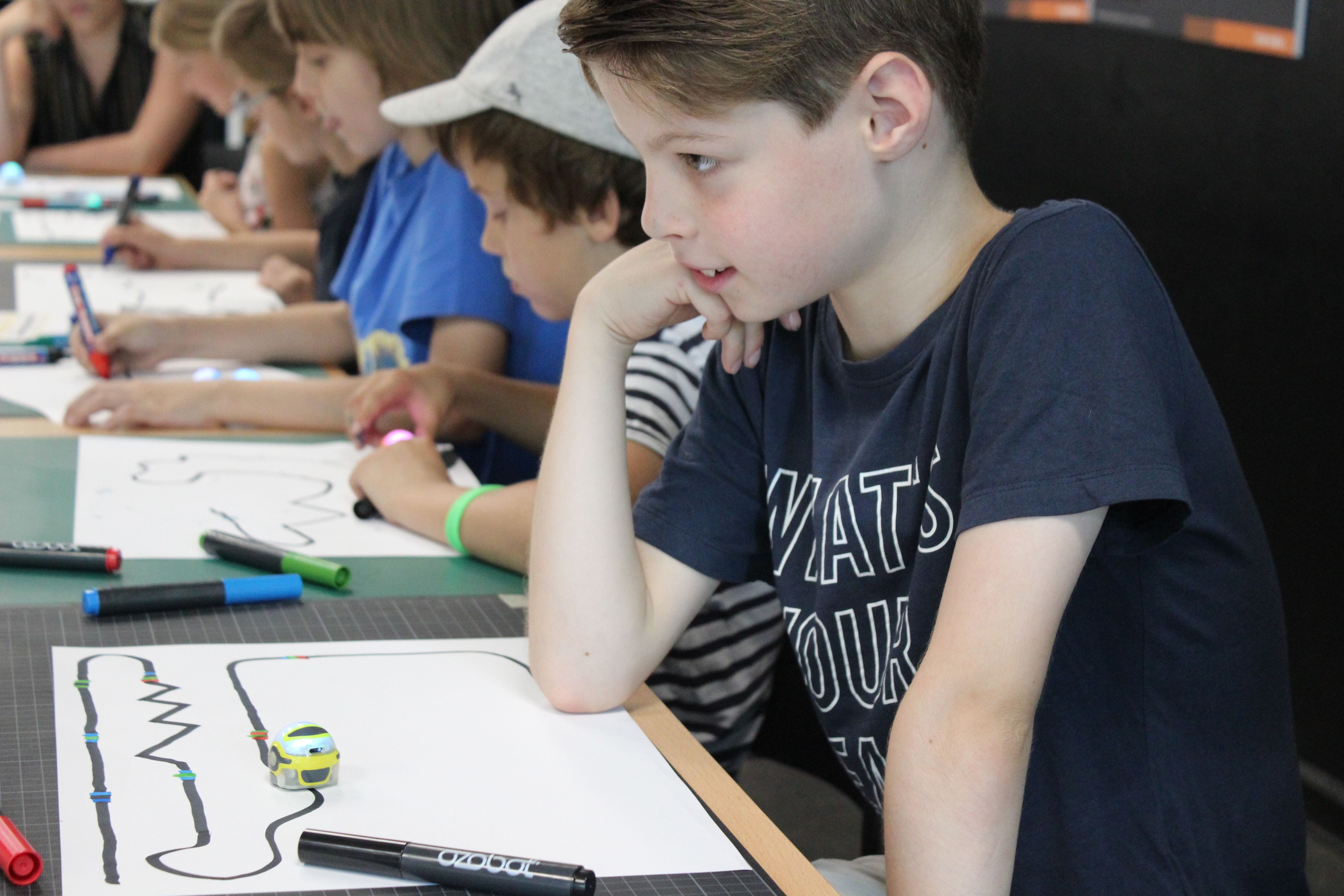 Junge schaut einem kleinen Roboter beim Fahren zu. Andere Kinder zeichnen Linien für Roboter.