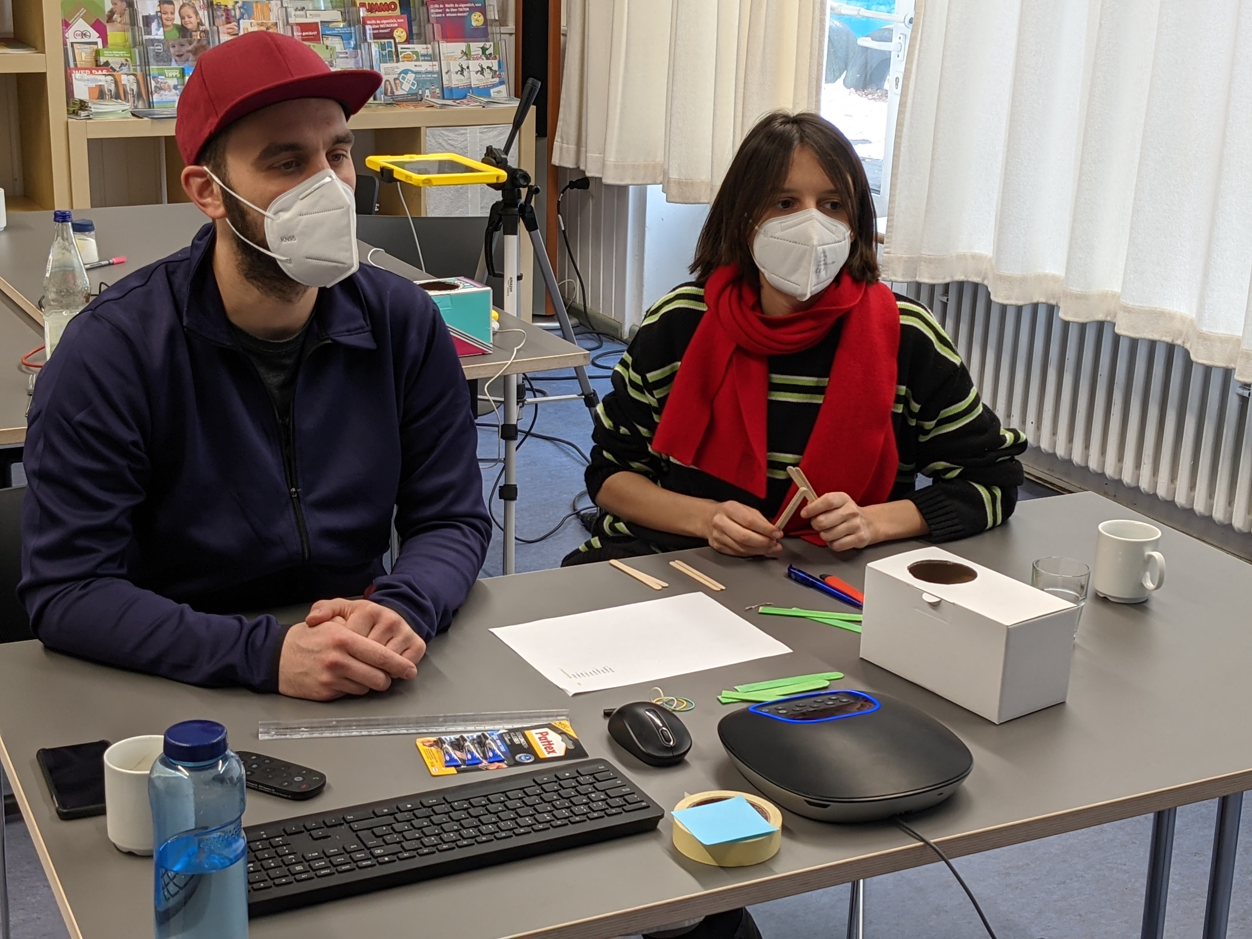 Zwei erwachsene Personen mit Maske sitzen an einem Tisch mit Technik